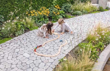 Zwei Kinder spielen mit einem Holzzug auf einem gepflasterten Gartenweg.