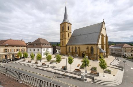 Eine Totale des Kirchenumfelds der St. Kilian Kirche in Scheßlitz. Im Vordergrund ist ein heller Platz mit Bäumen und Wasserbecken und im Hintergrund eine große Kirche zu sehen.