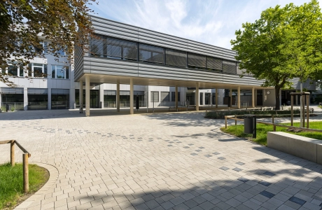 Offener Schulhof umgeben von grünen Wiesen, Bäumen und Sitzelementen. Ein Pfad bestehend aus anthrazitfarbenen Betonsteinen zieht sich von rechts nach links durch den gepflastertet Hof bis hin zu einem modernen, mehrteiligen Schulgebäude. Der vordere Teil steht auf Betonsäulen und ist dunkelgrau verkleidet. Der hintere Teil hat eine weiße Fassade. 