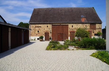 A rustic brick building with large wooden gates and a landscaped garden in the foreground. A paved path leads through the garden, which is planted with flowers and shrubs. In front of the garden is a natural beige paved courtyard laid out in rows. A garage with three large wooden doors can be seen on the left
