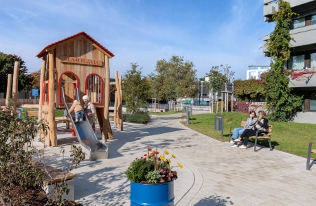 Spielplatzbereich mit einem großen Holzspielhaus und einer Metallrutsche, auf der sich zwei Personen befinden. Der umliegende Weg besteht aus beigen Pflastersteinen und führt durch eine begrünte Anlage. Rechts sitzen zwei weitere Personen auf einer Holzbank, im Vordergrund steht ein bepflanzter blauer Kübel, und im Hintergrund sind Bäume sowie moderne Gebäude zu sehen.
