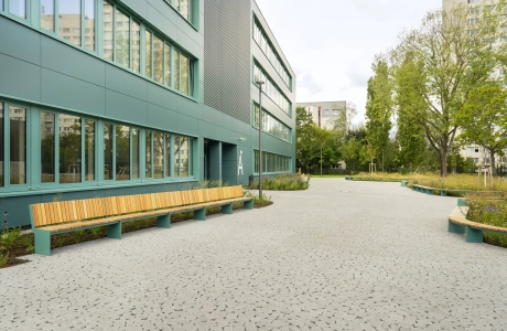 Ein mit rundlich natürlich wirkenden Betonsteinen gepflasterter Schulhof, welcher rechts neben einem modernen Schulgebäude mit petrolfarbener Fassade liegt. Um den gepflasterten Bereich ist alles mit Streuchern, Wiesen und Bäumen bepflanzt, weche mit Siztbänken eingerahmt werden.