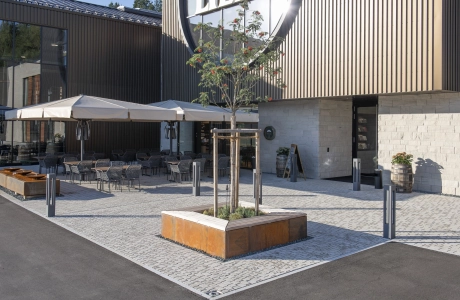 Moderner, gepflasterter Außenbereich mit Tischen und Stühlen unter weißen Sonnenschirmen vor einem modernem Gebäude mit Holz- und Steinfassade. Im Vordergrund ist ein kleinerer Baum, mit einer Einfassung aus rostigem Corthenstahl und Holzsitzfläche.