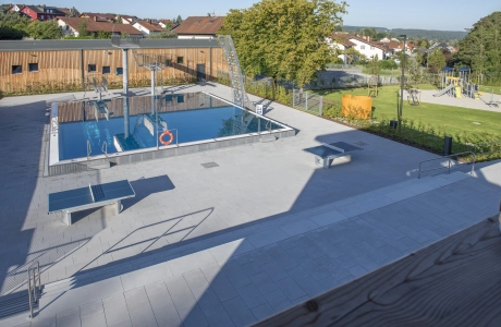Modernes Freibad mit rechteckigem Edelstahlbecken und weitläufiger, heller Terrasse mit mehreren Tischtennisplatten; im Hintergrund liegen eine Rasenfläche mit Bäumen und ein Spielplatz.