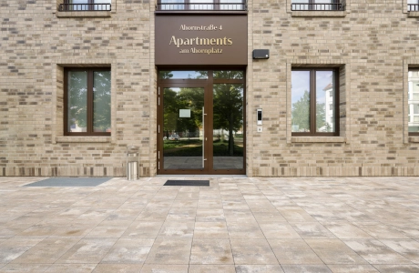 Gebäudefassade mit braunen Wandfliesen und dunkelbraunem Schild über der gläsernen Eingangstür. Davor ist ein Weg aus muschelkalkfarbenen Betonsteinen - dem GDM.KLIMASTEIN.