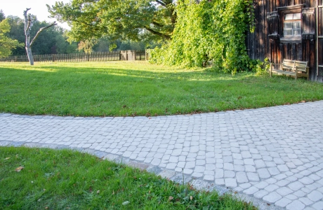 Geschwungener Gartenweg mit sandfarbenen Pflastersteinen, der entlang einer grünen Rasenfläche an einem mit Efeu bewachsenen Holzgebäude vorbeiführt, mit Bäumen, Holzzaun und Sitzbank in ruhiger, naturnaher Umgebung.