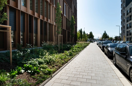 Weg aus Betonstein, der an einem Gebäude mit Grünstreifen entlang führt. Auf der anderen Seite des Weges parken Autos.