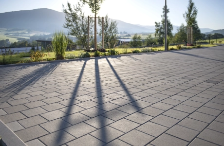 Nahaufnahme von grauem, versickerungsfähigem Betonsteinpflaster. Im Hintergrund ist eine hügelige Landschaft mit Sträuchern, Gräsern und Bäumen zu erkennen.