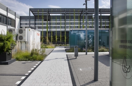Ein Grau gepflasterter Weg, der zu einem modernen Gebäude mit einer grün-bläulichen Fenster-Fassade führt. Auf dem Platz vor dem Gebäude sind Pflanzen, Container und Laternen zu erkennen.