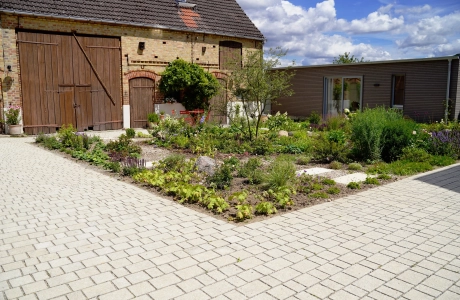 Ein rustikales Backsteingebäude mit großen Holztoren und einen angelegten Garten im Vordergrund. Ein gepflasterter Weg führt durch den Garten, der mit Blumen und Sträuchern bepflanzt ist. Vor dem Garten ist ein naturbeige gepflasterter Hof im Reihenverband angelegt. Rechts ist ein modernes Gebäude mit großen Glastüren zu sehen.