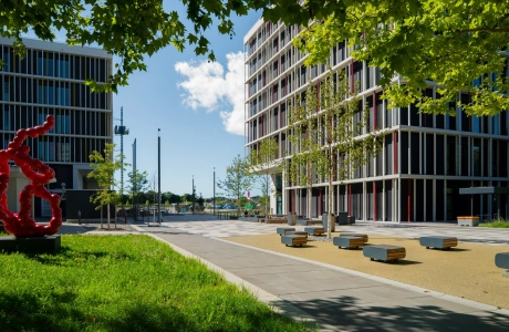 Moderner Universitätscampus mit mehrstöckigen Gebäuden, einer offenen Platzfläche mit Sitzbänken, jungen Bäumen und einer roten Skulptur, umgeben von Grün.