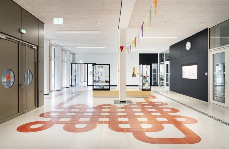 Der Eingangsbereich eines Schulgebäudes, durch dessen grau-beigen Schulboden sich ein rotes Band aus Sichtbeton-Platten zieht.