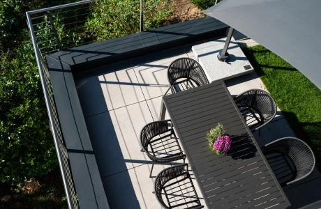 Draufsicht auf eine moderne Terrasse mit Betonsteinplatten. In der Mitte steht ein rechteckiger dunkelgrauer Outdoor-Tisch, umgeben von mehreren schwarzen Stühlen. Rechts ragt ein großer geöffneter Sonnenschirm über die Fläche. Die Terrasse ist von üppigem Grün und einem Geländer mit Edelstahlseilen eingerahmt.