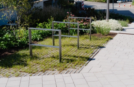 Anthrazitfarbene Fahrradständer auf bewachsenen Rasengittersteinen. Davor sind graue Pflastersteine verlegt. 