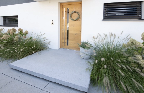 Moderner Hauseingang mit weißer Fassade, grauem Betonpodest und heller Haustüre aus Holz. Davor sind graue Betonsteinplatten verlegt. Links und Rechts von der Haustüre sind große, grüne Sträucher.