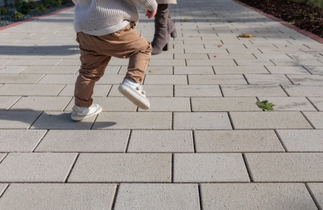Nahaufnahme eines Weges aus beigem GDM.KLIMASTEIN, auf dem ein Kind in Bewegung zu sehen ist. Im Hintergrund stehen weitere Personen entlang des Weges.