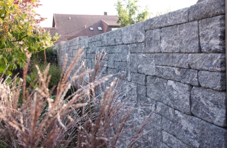 Lange, Grau-Schwarze GDM.DECA wall Sichtschutzmauer. Davor sind verschiedene Sträucher und Gräser zu finden.
