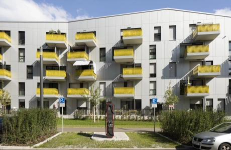 Mehrgeschossiges Wohngebäude mit gelben Balkonbrüstungen. Davor ist eine, mit GDM.SCADA liner, gepflasterte Parkfläche. Rechts und links neben der Fläche sind Beete mit grünen Sträuchern. Vor dem rechten Beet steht ein silbernes Auto.