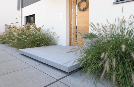 Moderner Hauseingang mit grauem Betonpodest und heller Haustüre aus Holz. Davor sind graue Betonsteinplatten verlegt. Links und Rechts von der Haustüre sind große, grüne Sträucher.