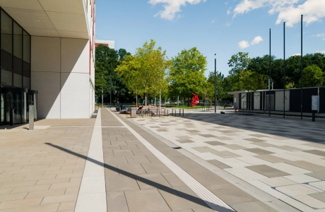 Weitläufiger Platz auf einem Universitätscampus entlang eines modernen Campusgebäudes mit Bäumen, Sitzgelegenheiten und einer roten Skulptur in der Ferne, gepflastert mit hellen und dunklen Betonsteinen.