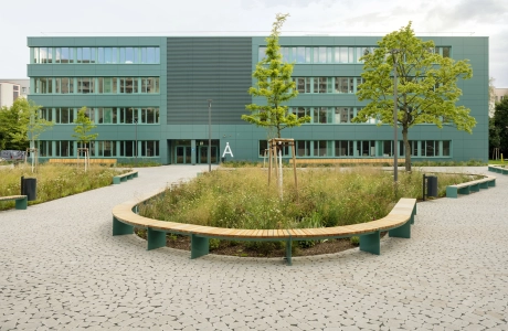 Ein mit rundlich gepflasterteten Betonsteinen gepflasterter Schulhof, welcher vor einem modernen Schulgebäude mit petrolfarbener Fassade liegt. Um den gepflasterten Bereich ist alles mit Streuchern, Wiesen und Bäumen bepflanzt, weche mit Siztbänken eingerahmt werden.