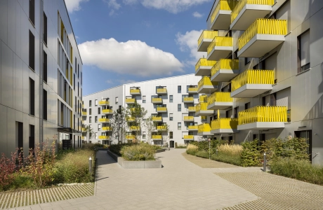 Blick auf einen großen Innenhof mit grün bewachsenen Beeten. Ringsherum sind mehrgeschossige Wohngebäude mit heller Fassade und gelben Balkonbrüstungen.