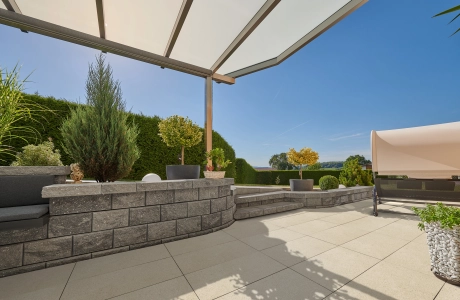 Eine Terrasse mit Dach, Mauer und Hecke im Hintergrund