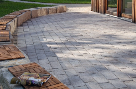 Ein großzügiger Terrassenbereich aus hellem GDM.CAMPA stone, eingefasst von einer niedrigen Steinmauer mit Holzauflagen, angrenzend an ein modernes Holzgebäude und eine grüne Rasenfläche.