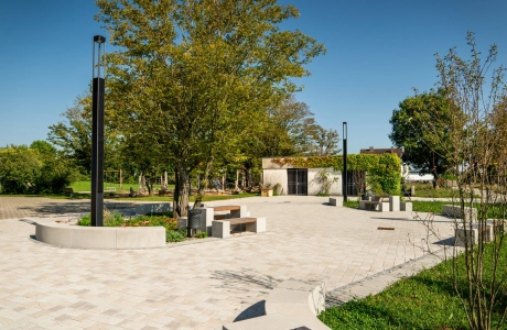 Kalk-Grau und Beige gepflasterter Schulhof mit vielen Sitzelementen und Grünflächen an einem sonnigen Tag mit blauem Himmel. Einige Bäume spenden Schatten. Im Hintergrund steht ein kleines weißes Gebäude, dessen Fassade mit Kletterpflanzen begrünt ist.