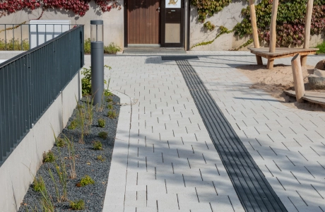 Gepflegter Außenbereich mit einem hellen, gepflasterten Weg. In den Weg ist ein durchgehendes Band aus anthrazitfarbenen Steinen integriert, das als Leitelement dient. Links verläuft ein bepflanztes Beet mit dunklem Splitt, rechts schließt ein Sandspielbereich mit Holzelementen an.