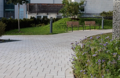 Blick auf einen geschwungenen Weg aus beigem GDM.KLIMASTEIN, der durch eine begrünte Außenanlage mit Blumenbeet, Bäumen und zwei Holzbänken führt. Im Hintergrund sind Gebäudefassaden und gepflegte Grünflächen sichtbar.