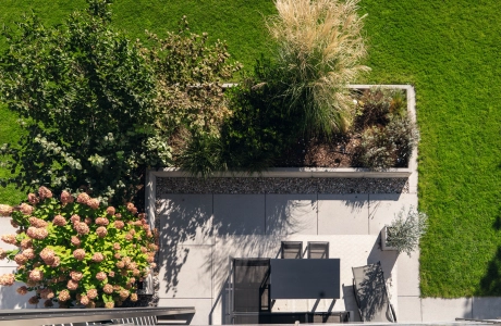 Draufsicht auf eine modern gestaltete Terrasse mit grauer GDM.NUEVA light. Die Fläche ist von üppigen Beeten mit grünen Sträuchern, Ziergräsern und blühenden Hortensien umrahmt. Auf der Terrasse steht ein schwarzer Tisch mit zwei Stühlen, angrenzend an eine gepflegte Rasenfläche.