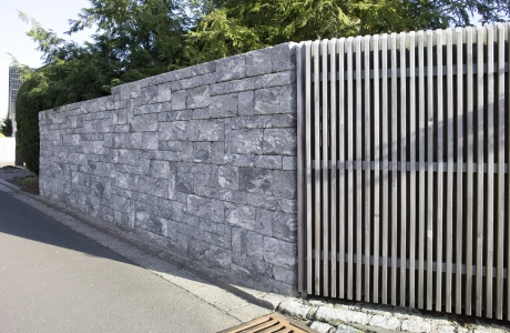 Sichtschutzmauer kombiniert aus GDM.DECA wall und Holzlatten. Davor ist eine geteerte Straße und hinter der Mauer sind Bäume und Sträucher zu erkennen.