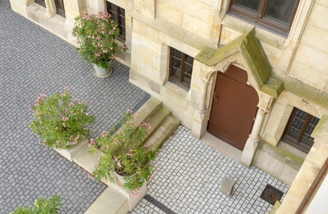 Entrance area of a historic villa with several storeys and elaborate architectural details. The building has a light-colored sandstone façade and ornate window frames. The anthracite and light gray paved area around the villa is well maintained and surrounded by planters.