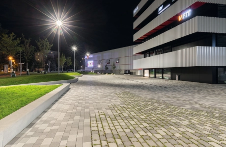 Gepflasterter Platz mit teilweise begrünter Fuge und an Grünfläche angrenzende Sitzmauern vor einem Sportzentrum