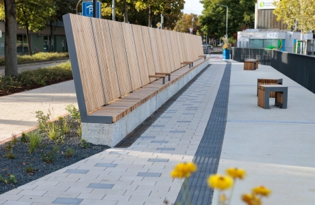 Promenade mit langen Holzsitzlehnen und einzelnen Sitzhockern, angelegt entlang eines Weges, der mit beigem GDM.KLIMASTEIN gepflastert ist; im Hintergrund Bäume, Straßenbereich und ein modernes Gebäude.