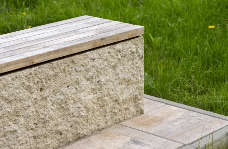 Moderne Sitzmöglichkeit aus dem kalksteinfarbenem GDM.MAXI block und hölzerner Sitzfläche. Der Sitzblock steht auf einer, aus Betonstein gepflasterten, Fläche. Außerdem befindet sich im Hintergrund eine Grünfläche