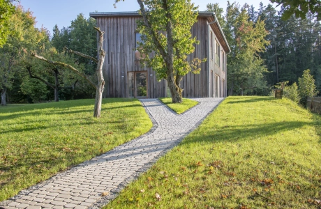 Mit GDM.BOLERO stone gepflasterter Gartenweg führt durch eine grüne Wiesenlandschaft zu einem modernen Holzhaus, umgeben von Bäumen und natürlicher Vegetation.