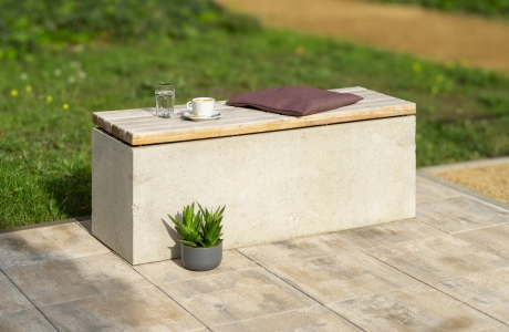 Moderne Sitzmöglichkeit aus dem kalksteinfarbenem GDM.MAXI block und hölzerner Sitzfläche. Darauf befinden sich ein Kissen, eine Tasse und ein Glas und vor der Sitzbank ist eine grüne Topfpflanze zu finden. Der Sitzblock steht auf einer, aus Betonstein gepflasterten, Fläche. 