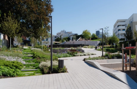 Ein granitfarbener Betonpflaster-Weg, der in einen Stadtgarten hineinführt. Links daneben befinden sich mit Grünpflanzen bewachsene Blockstufen. Im Stadtgarten findet man Sitzmöglichkeiten aus Beton und Holz, einen Spielplatz, ein Klettergerüst und Laternen.