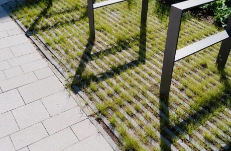 Nahaufnahme von anthrazitfarbene Fahrradständer auf bewachsenen Rasengittersteinen. Davor sind graue Pflastersteine verlegt. 