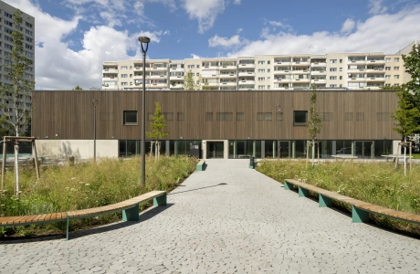 Frontalaufnahme eines großen Schulgebäudes mit Holzfassade mitten in einer Stadt. Vor dem Gebäude liegt ein Schulhof mit vielen Sitzmöglichkeiten, Tischtennisplatten und begrünten Flächen.