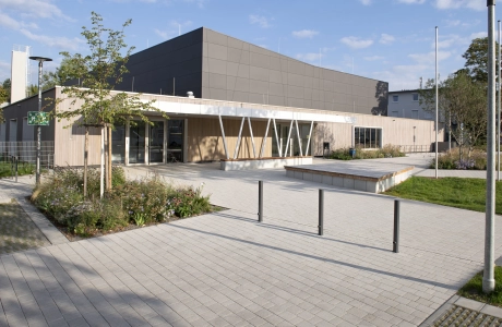 Moderne Außenfassade einer Sporthalle mit anthrazitfarbenen Platten im oberen und Holzverkleidung im unteren Bereich. Davor befindet sich ein mit Kalk-Grauen, versickerungsfähigen Betonsteinen gepflasterter Vorplatz mit Sträuchern, Gräsern und einem kleinen Baum.