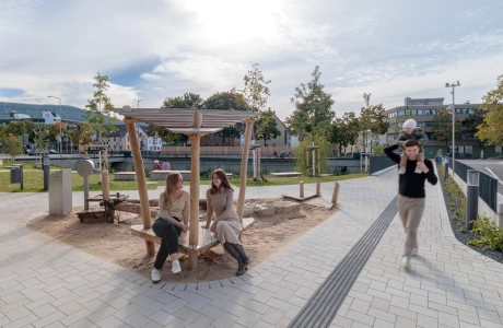 Außenbereich mit einem kleinen Spielplatz aus Holz sowie angrenzenden Grünflächen; zwei Personen sitzen am Rand des Sandspielbereichs, während eine weitere Person mit einem Kind vorbeigeht. Der umliegende Weg ist mit Betonsteinen gepflastert und führt durch die parkähnliche Anlage.