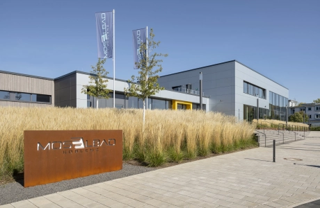 Im Vordergrund ist ein großes Schild aus Cortenstahl mit der Aufschrift "Moselbad Koblenz". Dahinter findet sich ein großes Beet mit beigem Ziergras. 