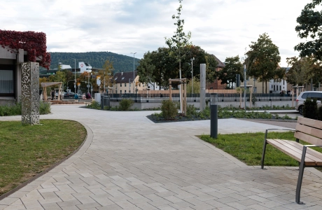 Geschwungener Weg aus hellem Betonstein, eingebettet in eine gepflegte Außenanlage mit jungen Bäumen, Beeten und Rasenflächen. Am Rand steht eine Holzbank, daneben befinden sich weitere Pflanzflächen und Gestaltungselemente.