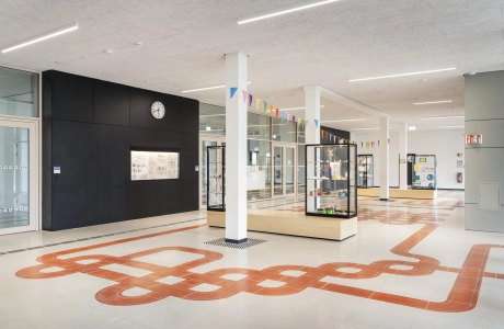 Der Eingangsbereich eines Schulgebäudes, durch dessen grau-beigen Schulboden sich ein rotes Band aus Sichtbeton-Platten zieht.