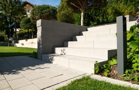 Breite Treppe aus GDM.NUEVA step, seitlich begrenzt durch die graue GDM.NOVO wall, eingebettet in eine offene Grünfläche mit Beeten und Gehölzern. Ein Weg, mit GDM.NUEVA stone gepflaster, führt auf die Treppe zu.