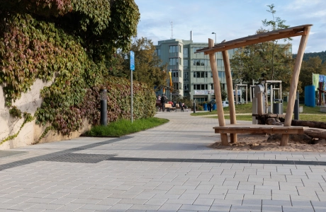 Außenbereich mit einem geschwungenen Weg aus beigem GDM.KLIMASTEIN, der an eine begrünte Mauer sowie an einen Sandspielbereich mit Holzelementen grenzt. Im Hintergrund befinden sich weitere Bereiche der Anlage mit Wegen, Pflanzen und Stadtmobiliar.