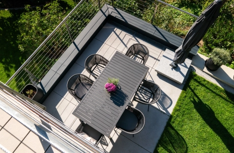 Draufsicht auf eine modern gestaltete Terrasse mit grauen GDM.NUEVA light Platten. Auf der Terrasse steht ein rechteckiger dunkelgrauer Tisch mit mehreren schwarzen Stühlen, daneben ein großer geschlossener Sonnenschirm. Die Fläche ist von Rasen sowie umliegenden Pflanzen eingerahmt und wird von einem Geländer mit Edelstahlseilen begrenzt.